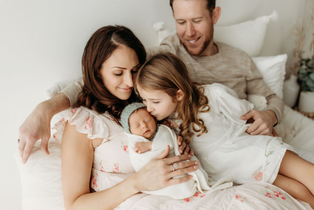 A family sits on a bed inside a beautiful natural light studio near Vancouver, Washington. They are admiring their newborn baby, who is being kissed on the cheek by her older sister.