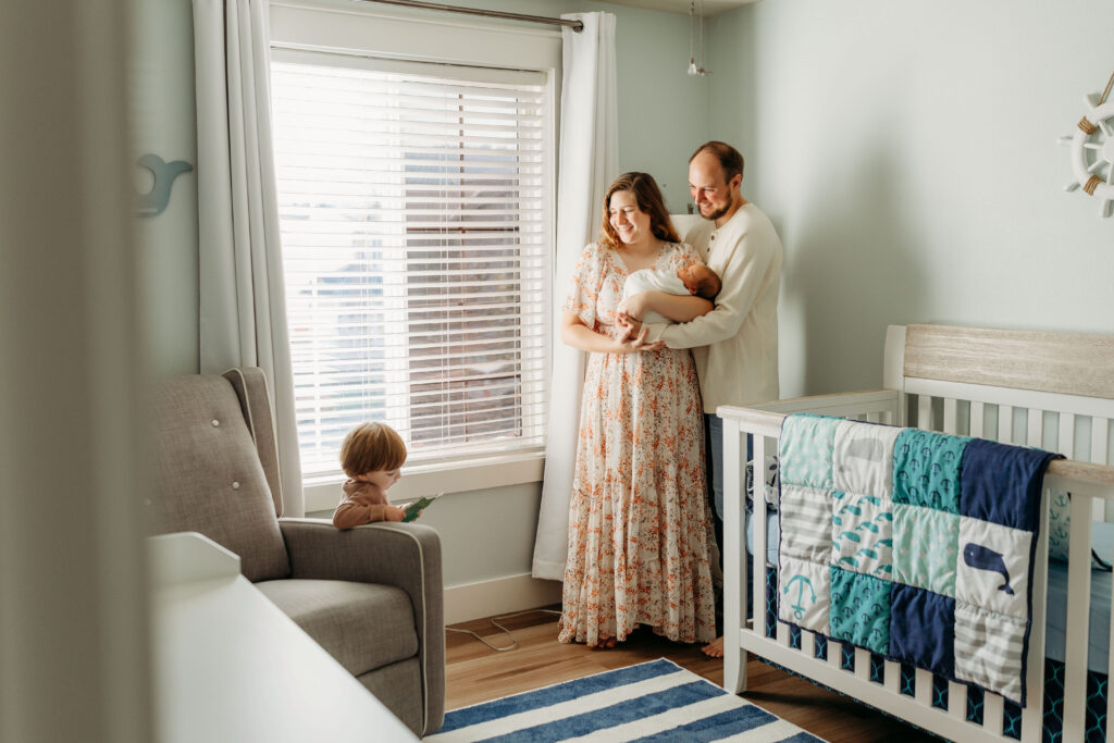 A couple stands in their baby's nursery, holding their newborn tenderly, while looking to their toddler who is reading a book across from them. The photo shows the intimacy of time shared by a family in the first days of welcoming a new baby.