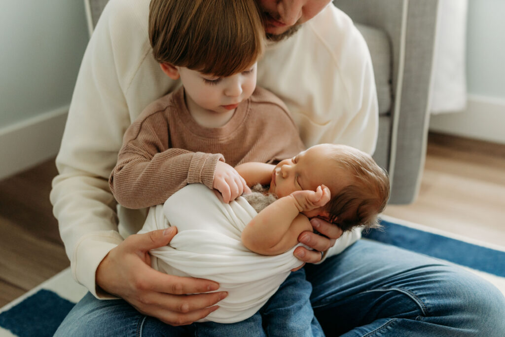 A toddler sits in his dad's lap wtih his newborn baby brother placed in front of him. He tenderly touches the newborn and examines his features. This photo shows the value of having a photographer capture your family with a newborn baby addition.