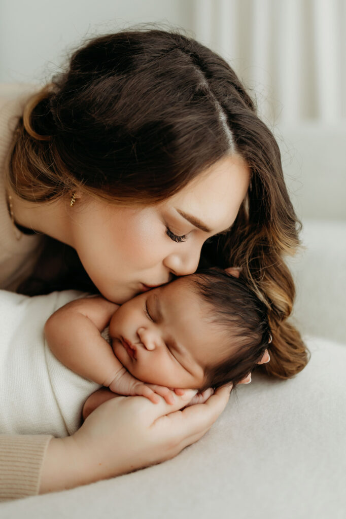 A mom kisses her newborn baby on the cheek. The baby's face is cradled in her hand, showing just how tiny she is compared to her mom. 