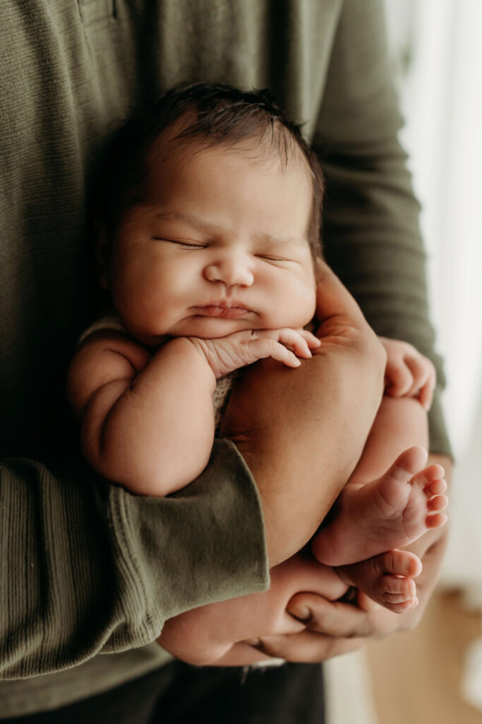 A newborn baby rests cradled in her dad's arms inside a Vancouver Washington natural light newborn studio. Dad's hand is across her chest and her arms and face are resting on it, showing how tiny she is in his arms.