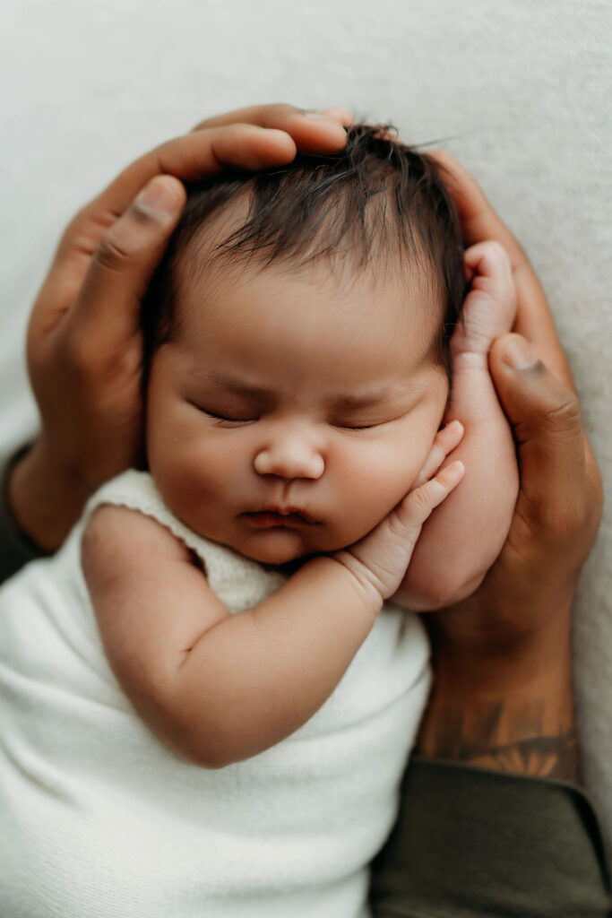 A newborn baby is photographed between her dad's two hands, showing just how small she is compared to him. 