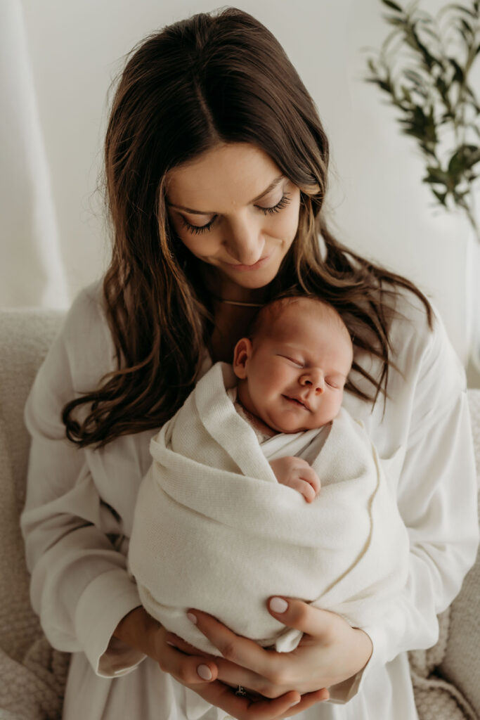 A newborn baby is shown being held by his mom as she looks down and admires him. He is smiling a content smile, showing the sweet connection between mom and baby in the early days. Photograph taken in a Vancouver WA area natural light newborn studio
