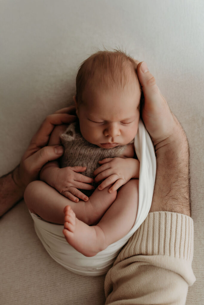 A newborn baby lays cradled in his dad's hands in a Vancouver WA area natural light newborn studio. The photo shows the cozy knit romper on the baby, which is available for use in the photographer's client wardrobe.