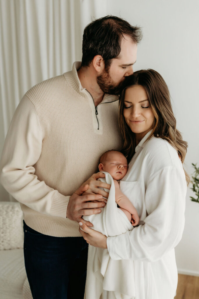 A mom holds her newborn baby on her chest, while her husband holds his hand on baby's back and leans in to kiss his wife on the head. An intimate, tender moment captured of the beginning of life as a family of three