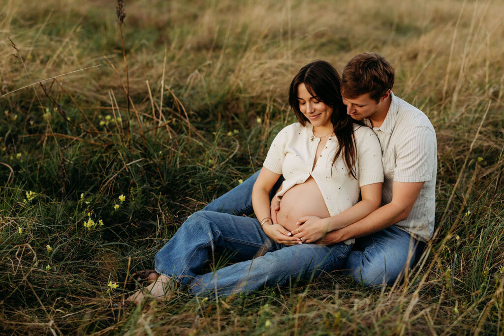 maternity couple sits in a field of flowers looking down at and rubbing mom's belly